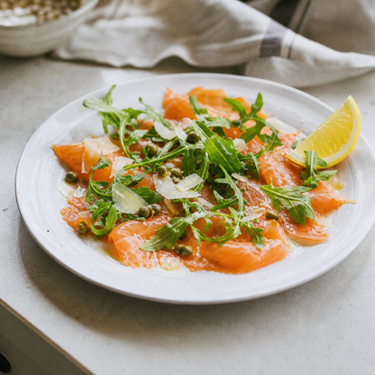 Gerookte zalm in lange plakken op een bord, met rucola, kappertjes en een citroenpartje